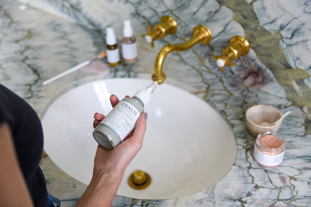 Alt Text: Oxalis Apothecary Facial Cleanser held over a marble sink, featuring purifying French Sea Clay and Pineapple Enzymes.
