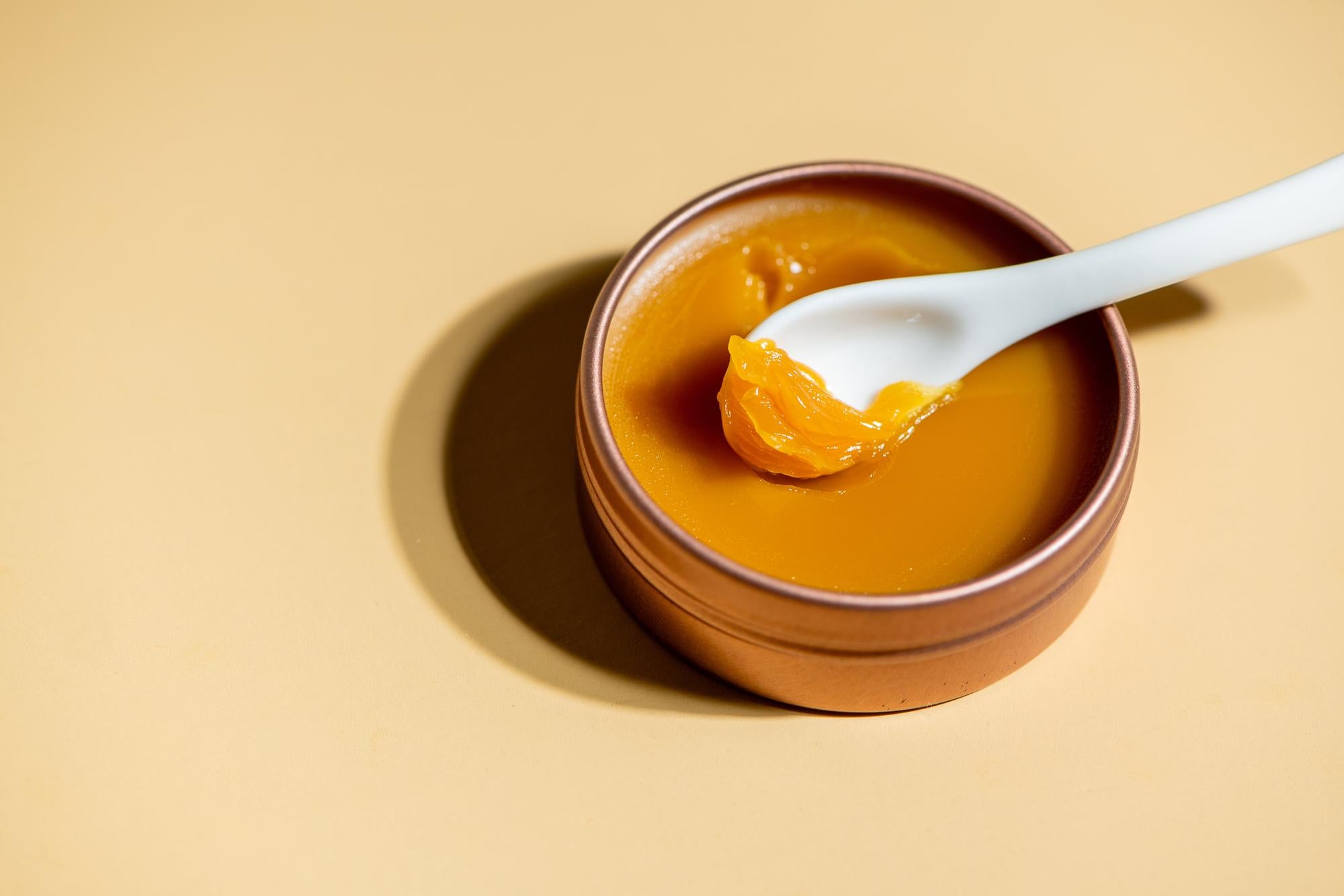 A top-down close-up of the Oxalis Apothecary Restorative Barrier Balm in a bronze tin. A white spatula scoops the rich, buttery yellow texture, highlighting the concentrated botanical formula for deep moisture barrier repair.