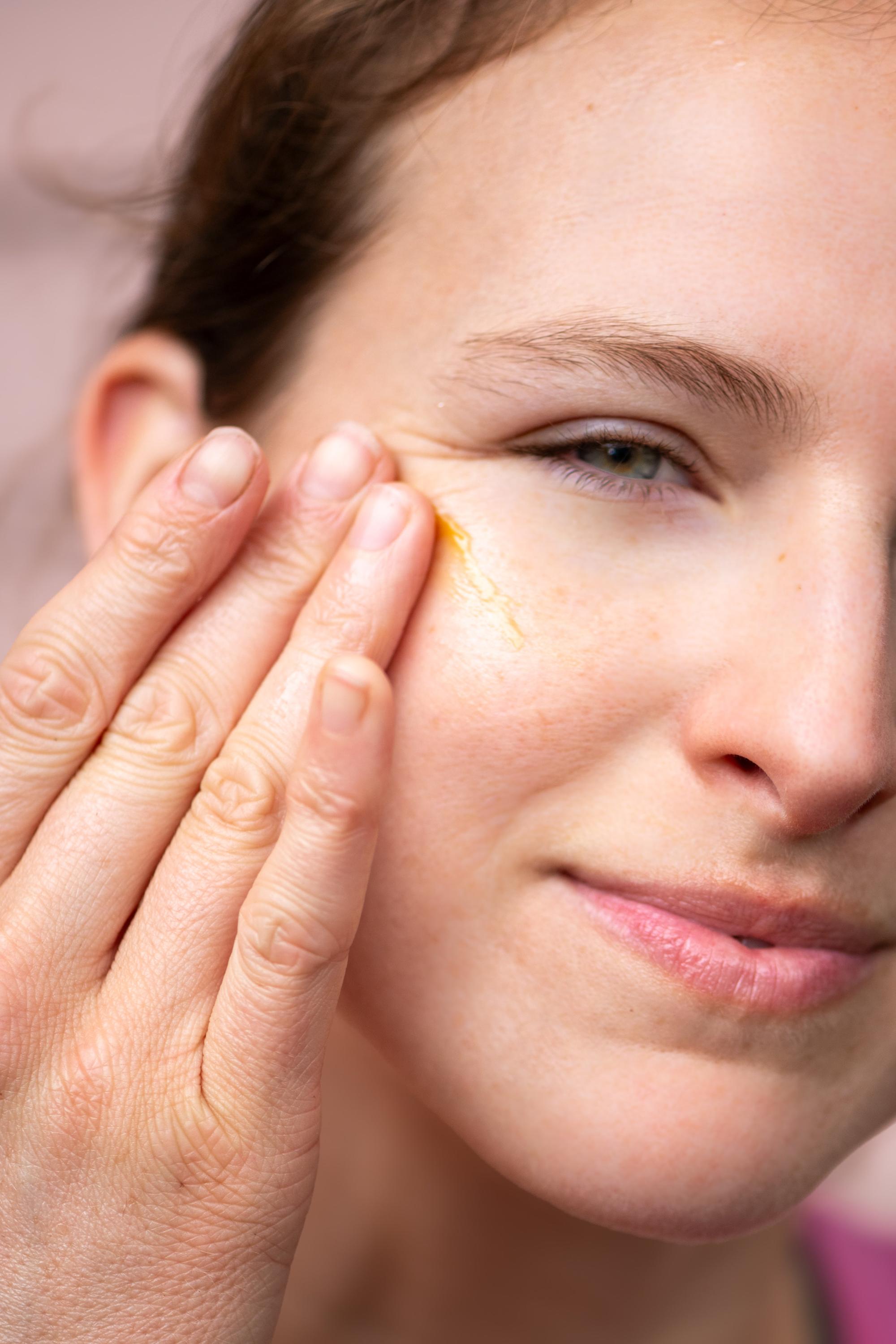 A close-up of a person applying the rich, yellow-toned Oxalis Apothecary Restorative Barrier Balm to their cheek to demonstrate the soothing texture and radiant glow it provides for moisture barrier repair.