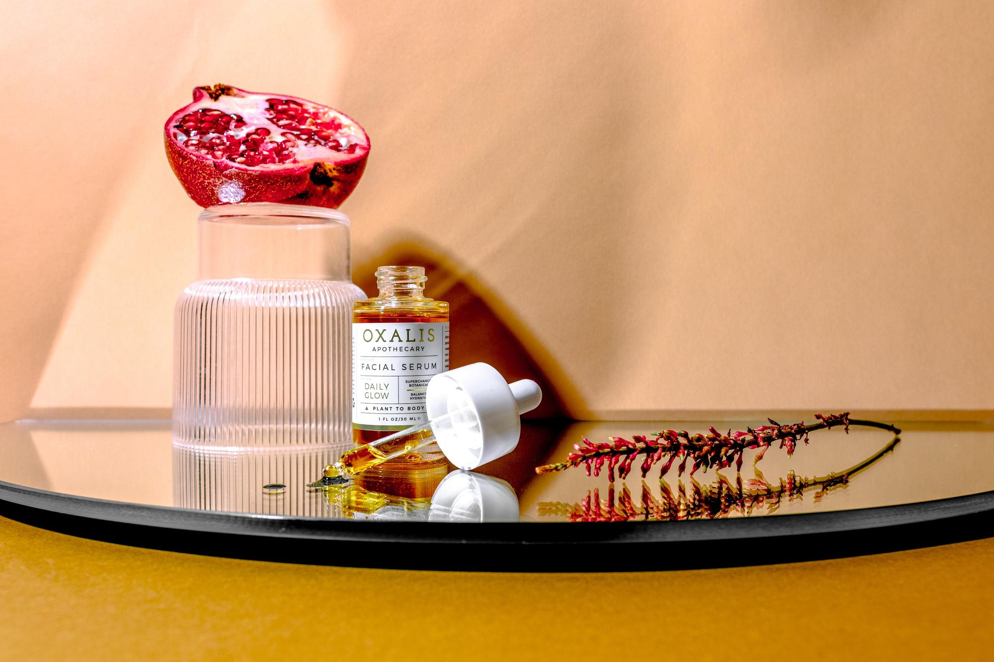 Oxalis Apothecary Daily Glow Facial Serum. A clear glass bottle of golden botanical oil with its dropper cap rests on a mirror base. Behind it, a halved pomegranate is displayed on a ribbed glass stand, representing its antioxidant-rich ingredients. Dried botanical accents surround the bottle in warm light.