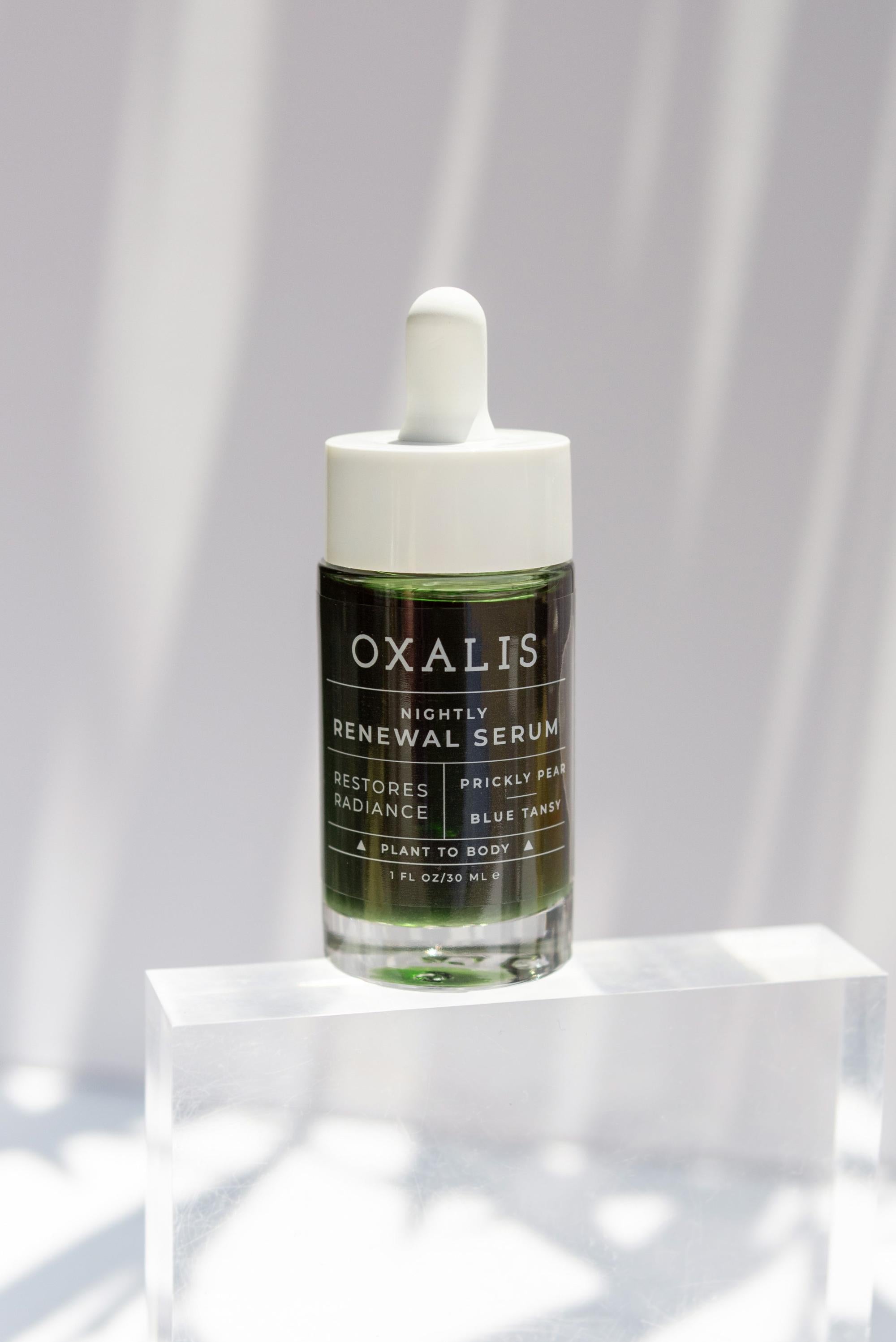 A bottle of Oxalis Apothecary Nightly Renewal Serum with Blue Tansy and Prickly Pear, positioned on a clear acrylic block under bright, dappled sunlight to highlight the rich green botanical formula.