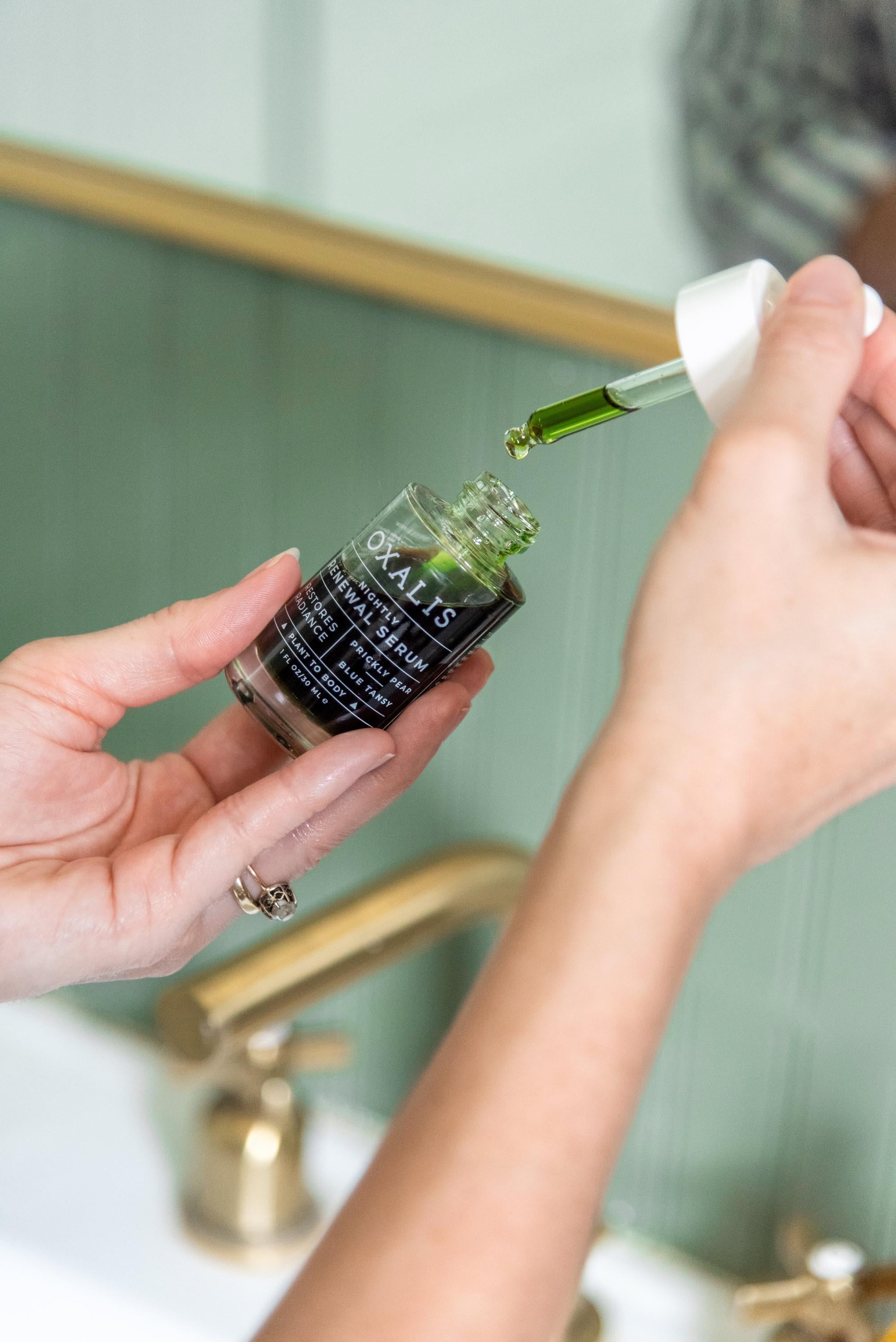 A close-up of hands using a glass dropper to apply the rich, dark green Oxalis Nightly Renewal Facial Serum, demonstrating the botanical oil texture and concentrated restorative formula.