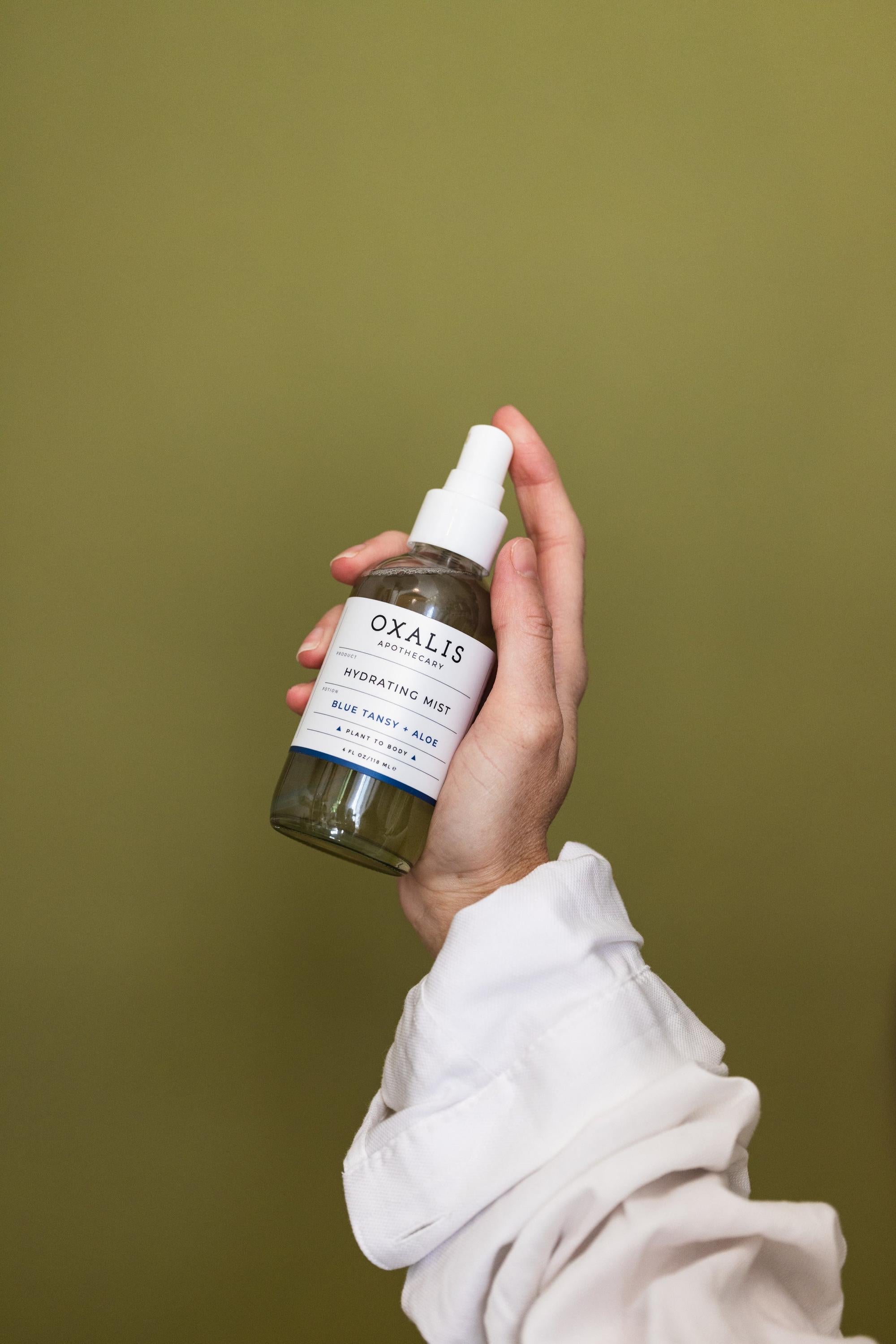 A hand in a white long-sleeved shirt holding a 4 oz bottle of Oxalis Apothecary Hydrating Mist against a solid olive green background. The label clearly displays 'Blue Tansy + Aloe' and the 'Plant to Body' branding.