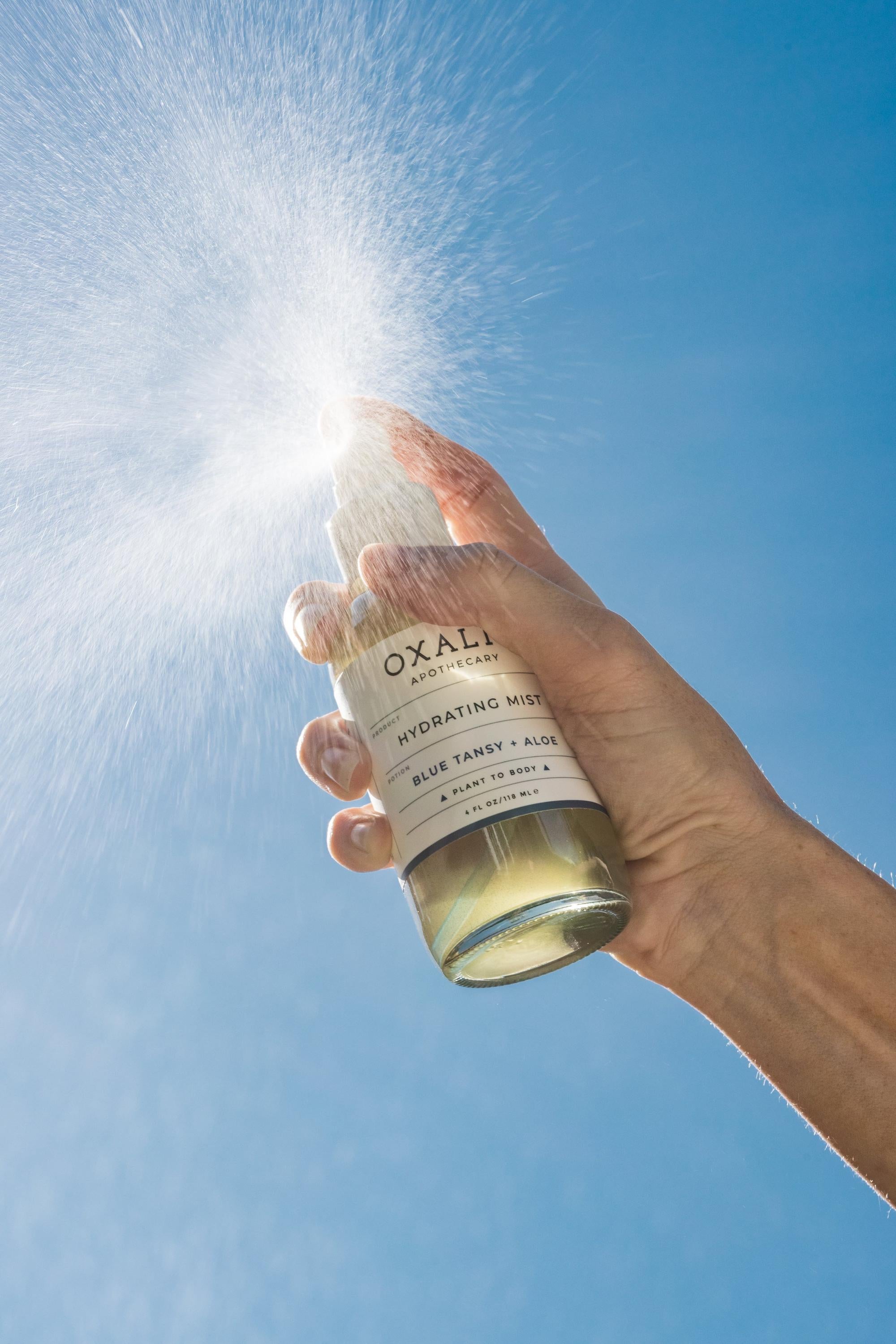 Action shot of Oxalis Apothecary Blue Tansy + Aloe Hydrating Mist being sprayed against a blue sky, showing the fine, refreshing mist of the botanical blue tansy hydrosol.