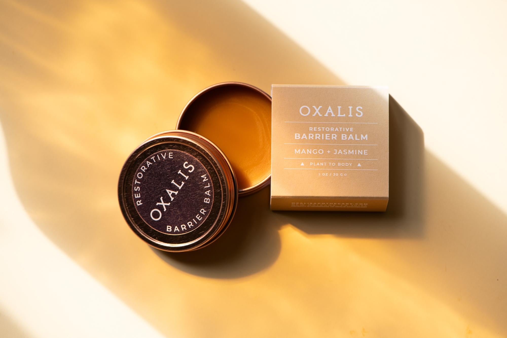 An open tin and box of Oxalis Apothecary Restorative Barrier Balm with Mango and Jasmine, styled in warm, natural sunlight to highlight the rich botanical ingredients for skin barrier repair.
