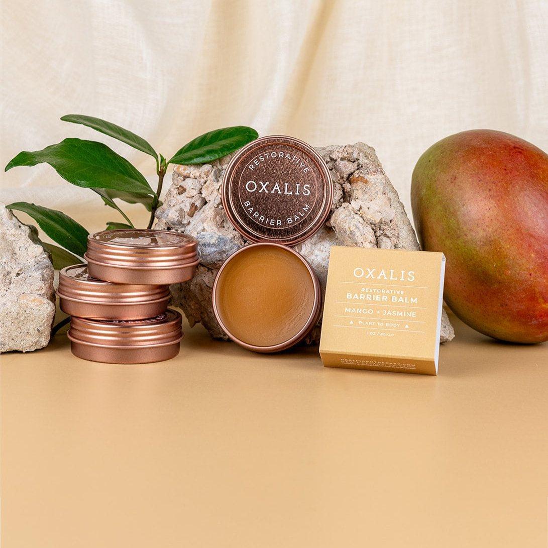 A stack of bronze tins and a box of Oxalis Apothecary Restorative Barrier Balm displayed with a fresh mango to highlight the soothing mango butter and jasmine ingredients for moisture barrier repair.