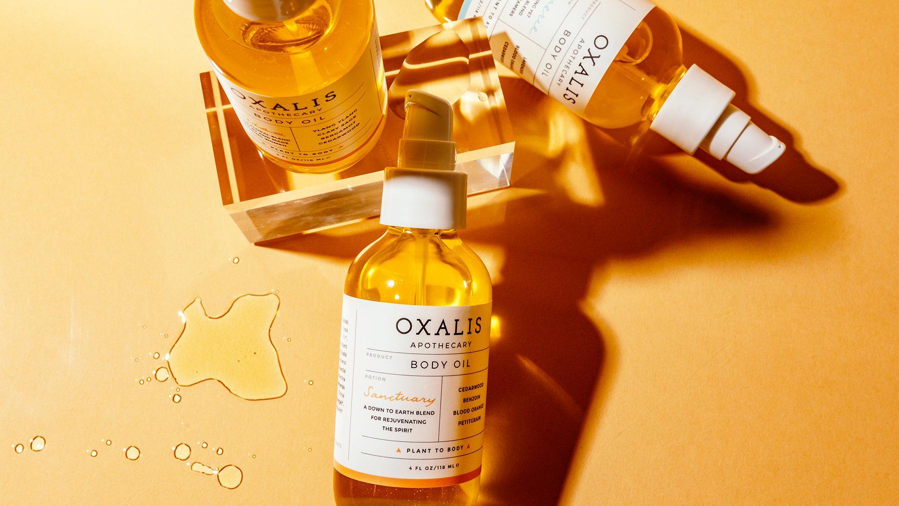 Top-down view of Oxalis Apothecary natural body oils including Sanctuary, Reverie, and Louisa, featuring golden botanical oil droplets on a warm, sunlit background.